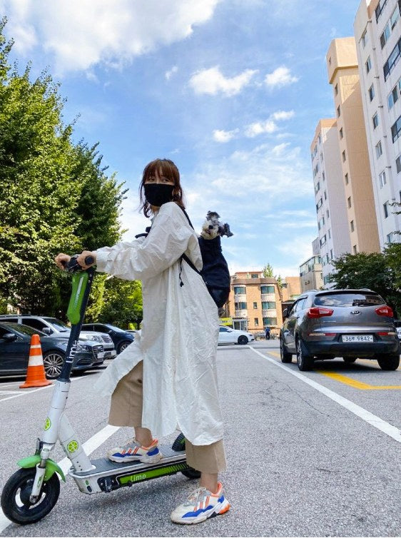 Woman riding a kick scooter on a city street with a dog carried on her back